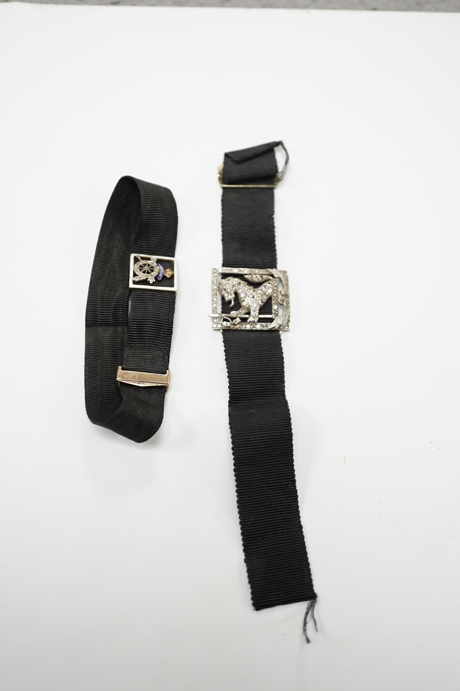 A black sash bracelet with diamond and enamel set Royal Fusiliers buckle, together with a similar sash with diamond set 'Charles' family crest, depicting a demi-rampant wolf, with motto.